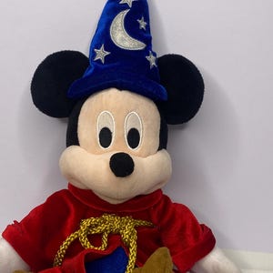 May include: A plush Mickey Mouse toy dressed as a sorcerer. Mickey wears a blue velvet wizard hat with silver crescent moon and star accents. He has a red velvet robe with a gold rope tie and brown shoes. The toy has black ears and a cream-colored face.