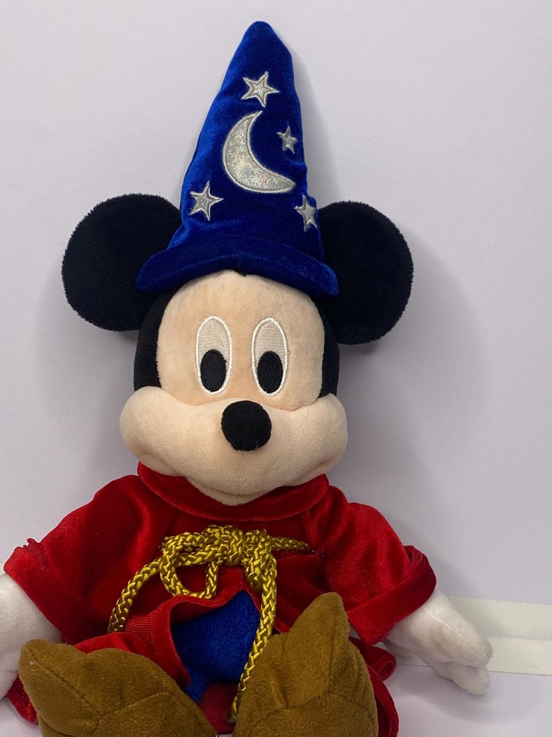 May include: A plush Mickey Mouse toy dressed as a sorcerer. Mickey wears a blue velvet wizard hat with silver crescent moon and star accents. He has a red velvet robe with a gold rope tie and brown shoes. The toy has black ears and a cream-colored face.