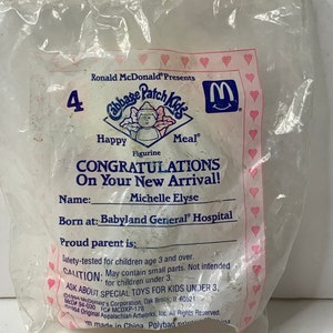Puede incluir: Una bolsa de plástico rosa y blanca con una figura de Cabbage Patch Kids en su interior. La bolsa tiene el logotipo de McDonald's y el texto "Happy Meal" y "¡Felicidades por tu nueva llegada!" La bolsa también incluye el nombre "Michelle Elyse" y el texto "Nacida en: Babyland General Hospital".