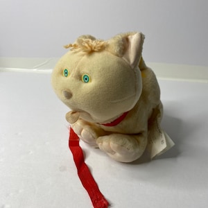 Puede incluir: Un juguete de peluche de gato de color crema con un collar rojo y ojos azules. El gato tiene una cinta roja larga atada a su pata.