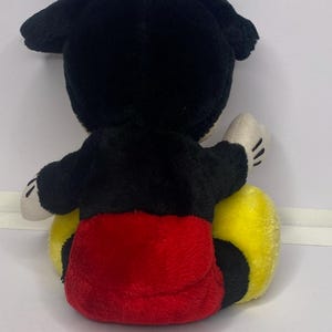 May include: A soft toy mouse, seen from behind. It has black ears and head, a black body, red shorts, and yellow feet. The hands are white with black lines for fingers.