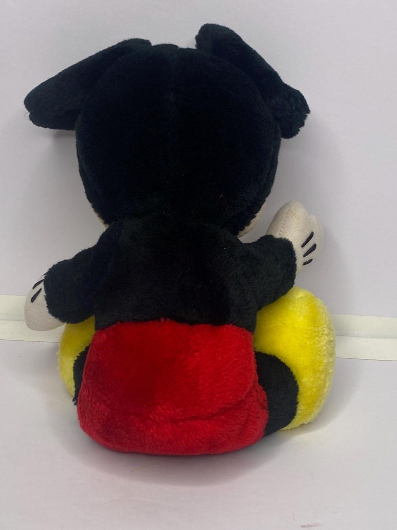 May include: A soft toy mouse, seen from behind. It has black ears and head, a black body, red shorts, and yellow feet. The hands are white with black lines for fingers.