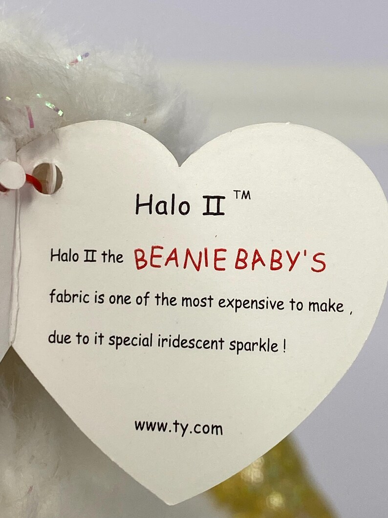 K&ouml;nnte beinhalten: Ein wei&szlig;es herzf&ouml;rmiges Etikett mit dem Text "Halo II" und "BEANIE BABY'S". Auf dem Etikett steht, dass der Stoff aufgrund seines schillernden Funkelns teuer ist. Die Website www.ty.com ist ebenfalls sichtbar.