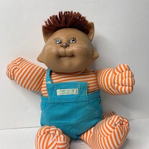 May include: A brown and white striped plush cat doll wearing blue overalls with a Cabbage Patch Kids logo. The doll has brown yarn hair and large blue eyes.