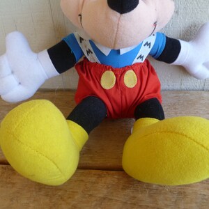Może przedstawiać: Pluszowa zabawka Mickey Mouse ubrana w czerwone spodenki z ż&oacute;łtymi guzikami i szelkami z nadrukowaną literą "M". Zabawka ma czarne buty z ż&oacute;łtymi podeszwami.