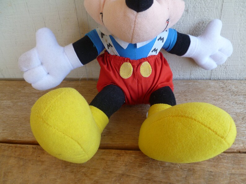 Może przedstawiać: Pluszowa zabawka Mickey Mouse ubrana w czerwone spodenki z ż&oacute;łtymi guzikami i szelkami z nadrukowaną literą "M". Zabawka ma czarne buty z ż&oacute;łtymi podeszwami.
