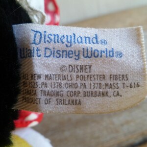 Op de afbeelding: Wit etiket met blauwe tekst met de tekst "Disneyland Walt Disney World &copy;DISNEY ALL NEW MATERIALS POLYESTER FIBERS 1525:PA 1378:OHIO PA 1378:MASS T-616 AHASA TRADING CORP. BURBANK, CA. PRODUCT OF SRILANKA"