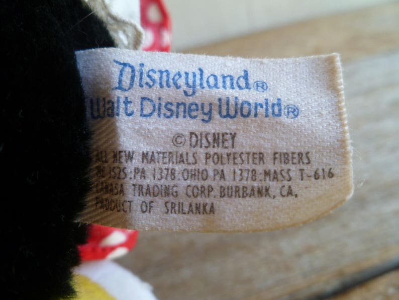 Op de afbeelding: Wit etiket met blauwe tekst met de tekst "Disneyland Walt Disney World &copy;DISNEY ALL NEW MATERIALS POLYESTER FIBERS 1525:PA 1378:OHIO PA 1378:MASS T-616 AHASA TRADING CORP. BURBANK, CA. PRODUCT OF SRILANKA"