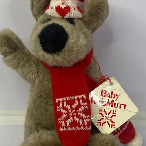May include: A soft brown dog toy, wearing a red knitted hat with white hearts and a red scarf with white snowflake patterns. The toy has a black nose and a tag that reads "Baby Kiss Mutt."