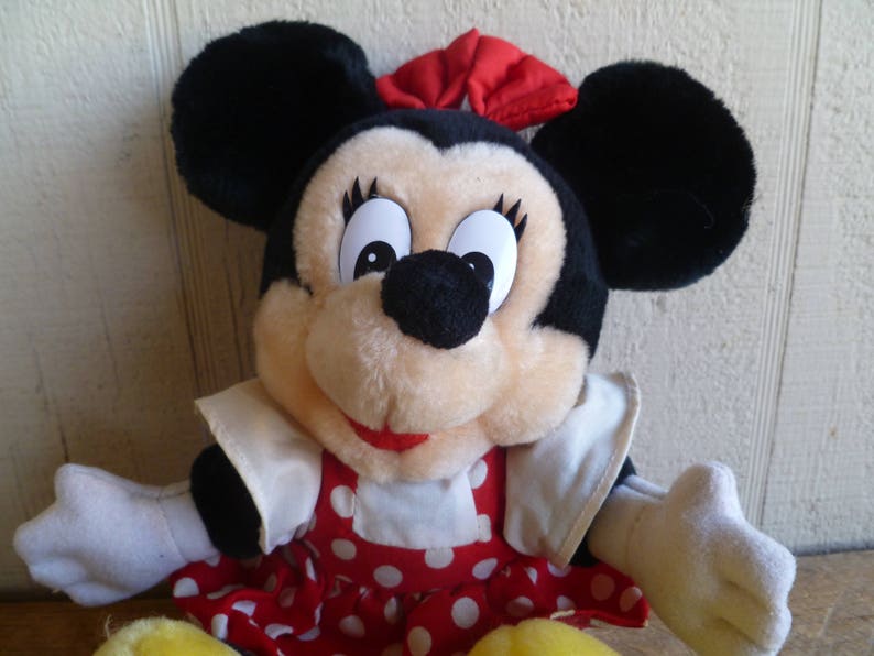 Op de afbeelding: Een pluche speelgoed van Mickey Mouse's vriendin Minnie Mouse. Ze draagt een rood-wit geblokt jurkje met een witte kraag en een rode strik. Ze heeft zwarte oren en een zwarte neus.