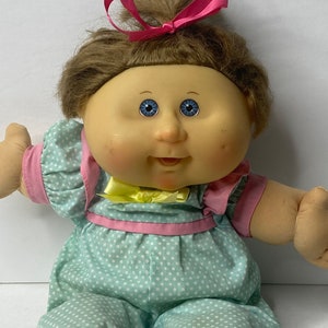 May include: A Cabbage Patch Kid doll with brown hair tied up with a pink ribbon. The doll is wearing a light blue and pink polka dot dress with a yellow bow.