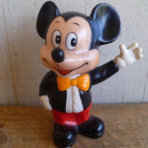Może przedstawiać: Plastikowa figurka Myszki Miki w kolorze czarno-białym, ubrana w czarny smoking z pomarańczową muszką. Figurka ma czerwono-białe ciało i podnosi prawą rękę.