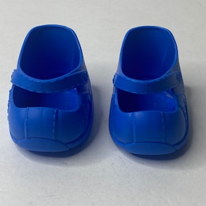 Może przedstawiać: Para niebieskich plastikowych butów dla lalki z paskiem na górze.
