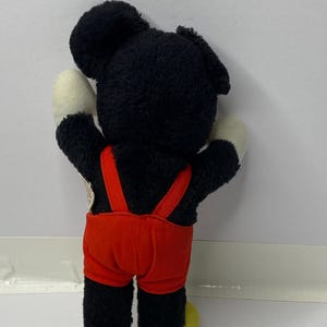 May include: Black and red plush Mickey Mouse toy with yellow shoes. The toy is wearing red overalls.