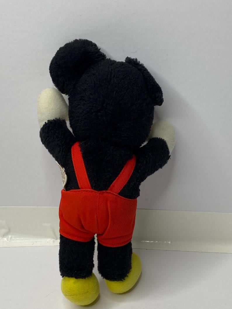 May include: Black and red plush Mickey Mouse toy with yellow shoes. The toy is wearing red overalls.