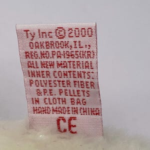 K&ouml;nnte beinhalten: Nahaufnahme eines rechteckigen Etiketts mit rotem Text auf wei&szlig;em Hintergrund. Auf dem Etikett steht "Ty Inc. &copy; 2000" und weitere Details. Das Etikett ist an einem Pl&uuml;schtier befestigt.