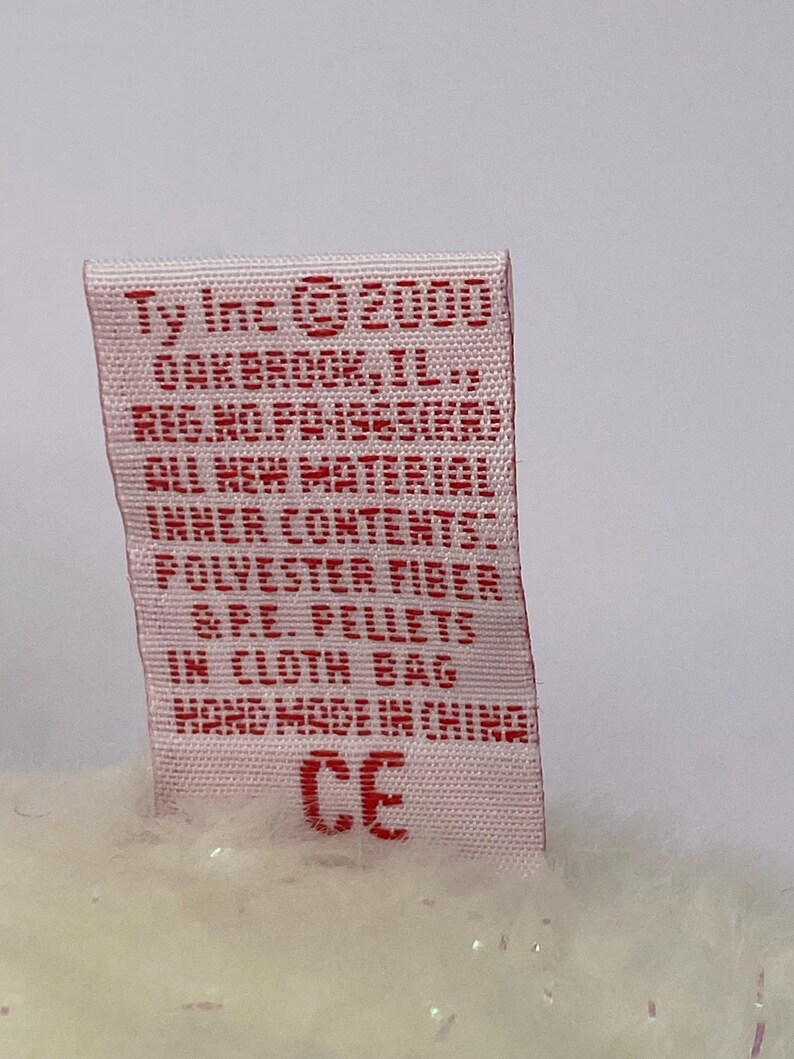 K&ouml;nnte beinhalten: Nahaufnahme eines rechteckigen Etiketts mit rotem Text auf wei&szlig;em Hintergrund. Auf dem Etikett steht "Ty Inc. &copy; 2000" und weitere Details. Das Etikett ist an einem Pl&uuml;schtier befestigt.