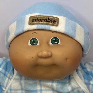 Puede incluir: Una muñeca vintage con una cara redonda, ojos verdes y una nariz pequeña. La muñeca lleva un gorro a rayas azules y blancas con la palabra "adorable" en un parche. El atuendo de la muñeca es a cuadros azules y blancos.