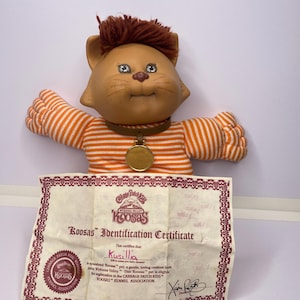 May include: A vintage Koosas doll, featuring a cat-like face with brown hair and blue eyes. It wears an orange and white striped shirt and a brown collar with a gold medallion. An identification certificate is included.