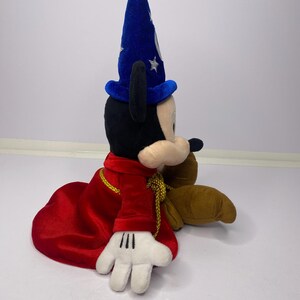 May include: A plush Mickey Mouse toy dressed as a sorcerer, wearing a blue hat with silver stars and a red robe with gold trim. The toy has black ears, a black nose, and white gloves.
