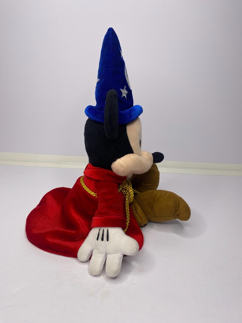 May include: A plush Mickey Mouse toy dressed as a sorcerer, wearing a blue hat with silver stars and a red robe with gold trim. The toy has black ears, a black nose, and white gloves.