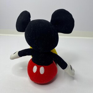 May include: A black and red plush Mickey Mouse toy with yellow accents. The toy is sitting on a white surface.