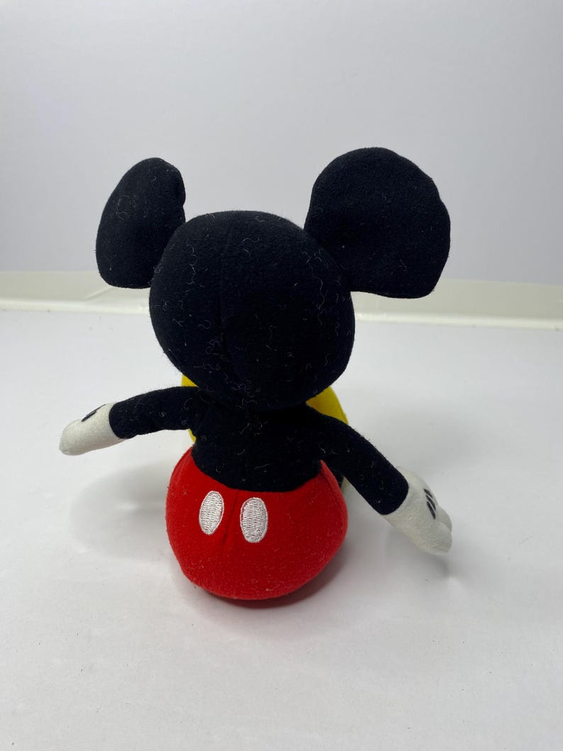 May include: A black and red plush Mickey Mouse toy with yellow accents. The toy is sitting on a white surface.