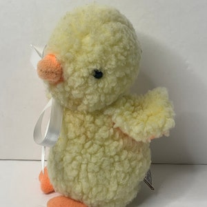 May include: A yellow plush chick with a white bow and orange feet. The chick is made of fluffy material and has a sweet expression.