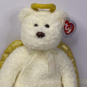 K&ouml;nnte beinhalten: Ein wei&szlig;er Teddyb&auml;r mit goldenem Heiligenschein und Fl&uuml;geln. Der B&auml;r hat eine cremefarbene Schleife und ein rotes herzf&ouml;rmiges Etikett. Ein Sammlerst&uuml;ck.