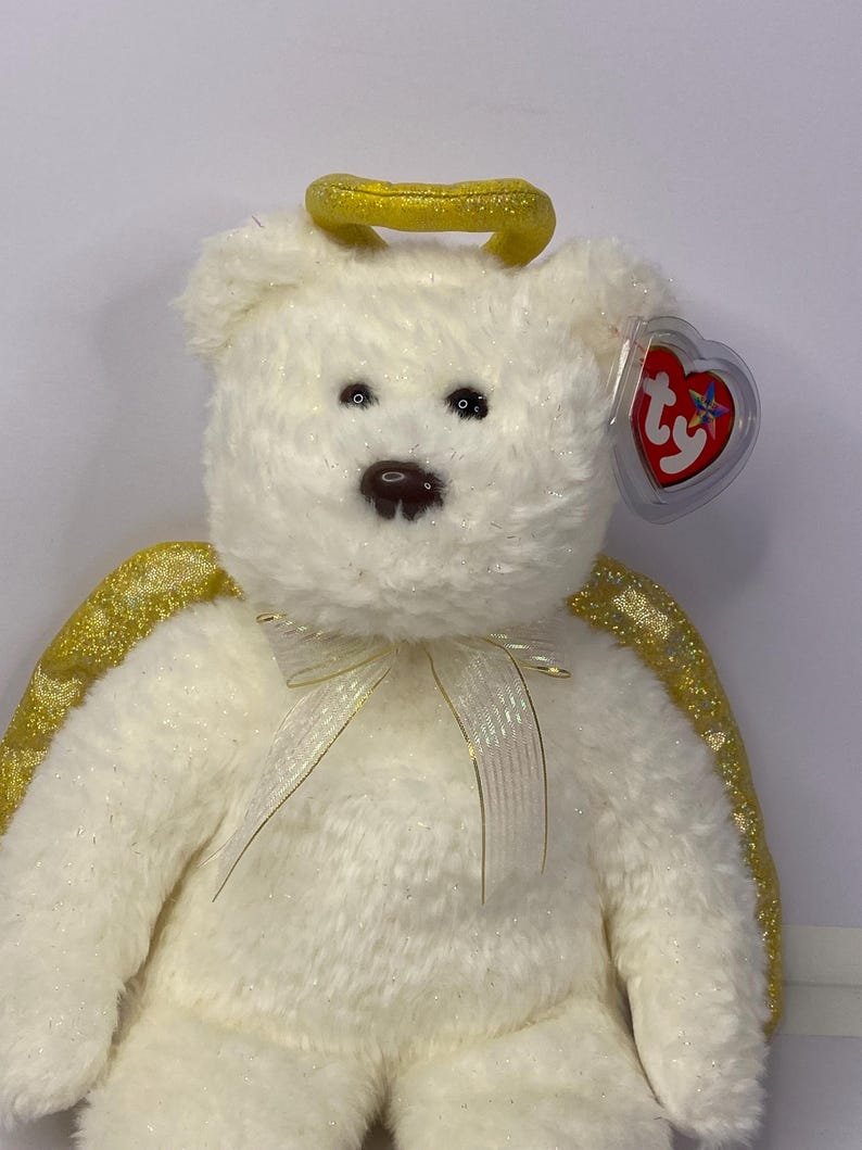 K&ouml;nnte beinhalten: Ein wei&szlig;er Teddyb&auml;r mit goldenem Heiligenschein und Fl&uuml;geln. Der B&auml;r hat eine cremefarbene Schleife und ein rotes herzf&ouml;rmiges Etikett. Ein Sammlerst&uuml;ck.