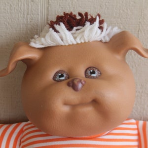 May include: A brown plush toy with a white and brown yarn hairdo. The toy has brown eyes, a brown nose, and a smiling mouth. The toy is wearing an orange and white striped shirt.