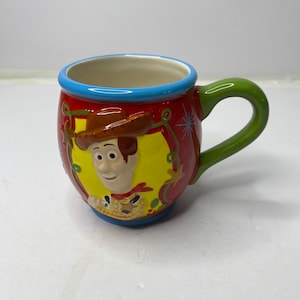 Puede incluir: Una taza de cer&aacute;mica con una imagen de dibujos animados de Woody de Toy Story. La taza tiene un fondo rojo con un &oacute;valo amarillo que enmarca la cara de Woody. El asa es verde y el borde es azul. La taza est&aacute; dise&ntilde;ada para bebidas.