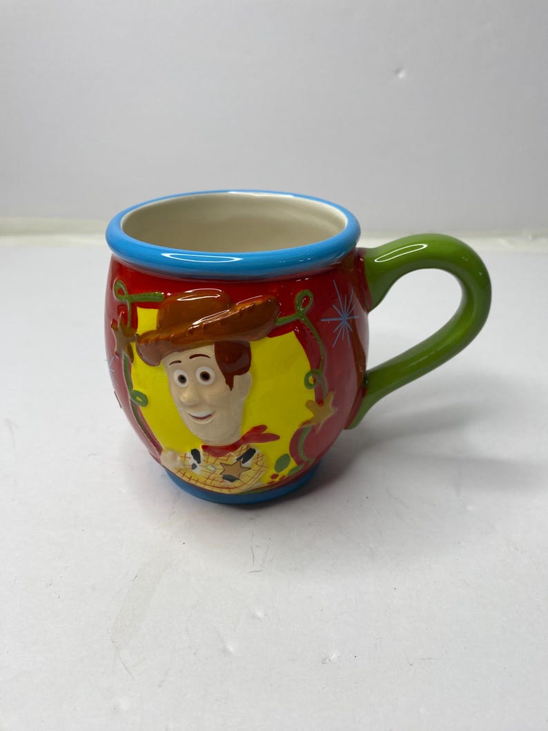 Puede incluir: Una taza de cer&aacute;mica con una imagen de dibujos animados de Woody de Toy Story. La taza tiene un fondo rojo con un &oacute;valo amarillo que enmarca la cara de Woody. El asa es verde y el borde es azul. La taza est&aacute; dise&ntilde;ada para bebidas.