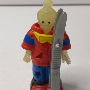May include: A red, blue, and yellow plastic figurine of a person wearing a ski jacket and holding a gray ski. The figurine has a translucent head with a yellow bun.