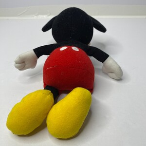 May include: A plush Mickey Mouse toy with black fur, red shorts, and yellow shoes. The toy is lying on its back with its arms outstretched.