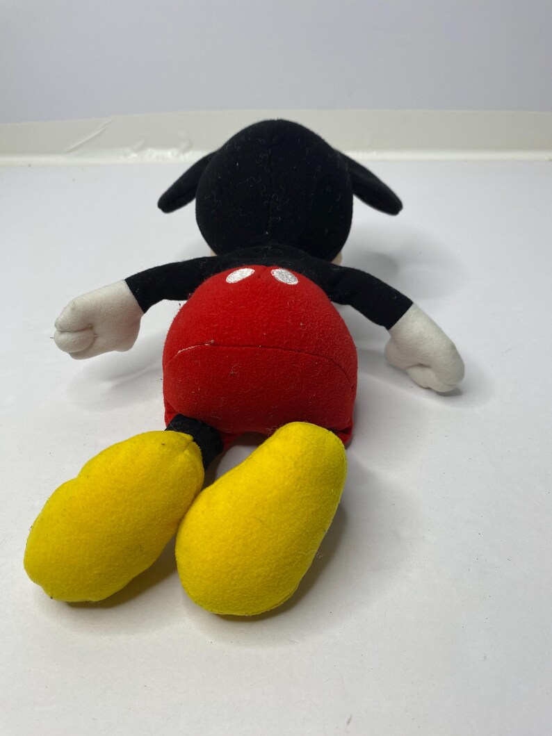 May include: A plush Mickey Mouse toy with black fur, red shorts, and yellow shoes. The toy is lying on its back with its arms outstretched.