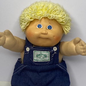 Peut inclure: Une poupée vintage Cabbage Patch Kids avec un teint clair, des yeux bleus et une coiffure en fil jaune. La poupée porte une salopette bleu denim avec des boutons blancs et un écusson vert et blanc avec l'inscription "Cabbage Patch Kids".