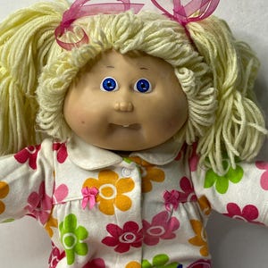 Puede incluir: Una muñeca Cabbage Patch Kid con pelo de lana rubio, ojos azules y un lazo rosa. La muñeca lleva un mono blanco con estampado floral.