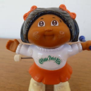 May include: A vintage Cabbage Patch Kids doll figurine. The doll has dark hair with orange bows, brown skin, and is wearing a white shirt with an orange skirt. It is holding blue and white batons.