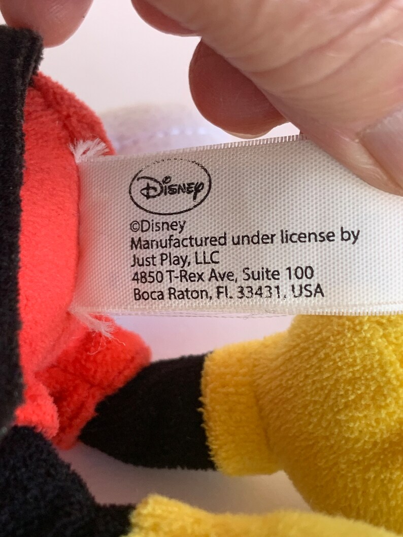Op de afbeelding: Een close-up van een Disney-productlabel. Het label is wit met het Disney-logo en de tekst "&copy;Disney Vervaardigd onder licentie door Just Play, LLC 4850 T-Rex Ave, Suite 100 Boca Raton, Fl. 33431, USA."