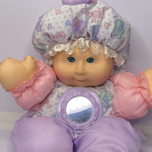 May include: A vintage baby doll with a soft, plush body and a round, smiling face. The doll wears a patterned bonnet, a dress with a mirror, and has pink and lavender accents. The doll's eyes are green.