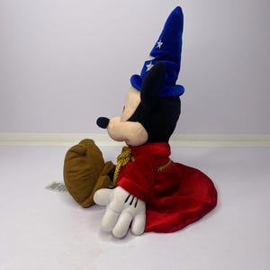 May include: A plush Mickey Mouse toy dressed as a sorcerer, wearing a blue hat with white stars and a red robe. The toy has black ears, a black nose, and white gloves. The plush is sitting down.
