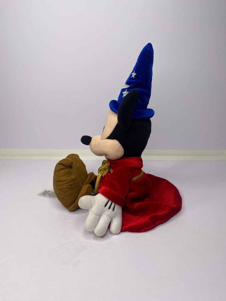 May include: A plush Mickey Mouse toy dressed as a sorcerer, wearing a blue hat with white stars and a red robe. The toy has black ears, a black nose, and white gloves. The plush is sitting down.