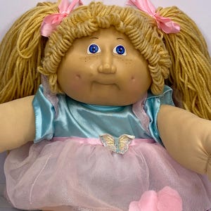 May include: A vintage doll with blonde yarn hair styled in pigtails, each tied with a pink ribbon. The doll has blue eyes, freckles, and a light blue satin top with a pink tulle skirt. A butterfly embellishment adorns the waist.
