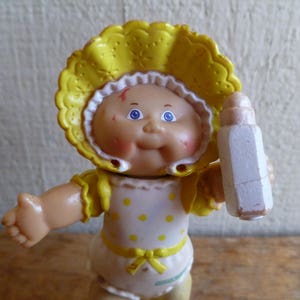 May include: A yellow and white plastic doll with a yellow bonnet and a white dress with yellow polka dots. The doll is holding a white plastic bottle.