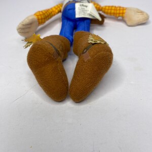 May include: A plush toy of a cowboy character, featuring a brown hat, yellow plaid shirt, blue pants, and brown boots. The toy has a white tag and is lying on a white surface. The toy's arms and legs are outstretched.
