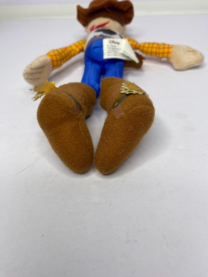 May include: A plush toy of a cowboy character, featuring a brown hat, yellow plaid shirt, blue pants, and brown boots. The toy has a white tag and is lying on a white surface. The toy's arms and legs are outstretched.