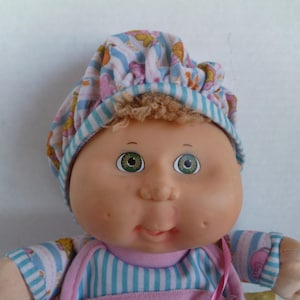 May include: A Cabbage Patch Kids doll with green eyes and a blue and white striped dress. The doll is wearing a pink and white patterned bonnet.