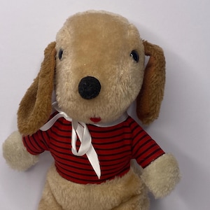 Puede incluir: Un peluche de perro con pelaje marrón claro, orejas marrones y una nariz negra. Viste una camiseta a rayas rojas y negras con ribete blanco y una cinta blanca. El juguete tiene patas blancas y una pequeña etiqueta.