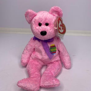 May include: A pink plush bear with a purple ribbon and an Easter egg applique. The bear has black eyes and a black nose. A red and white tag is attached to the bear's ear. The bear is sitting on a white surface.